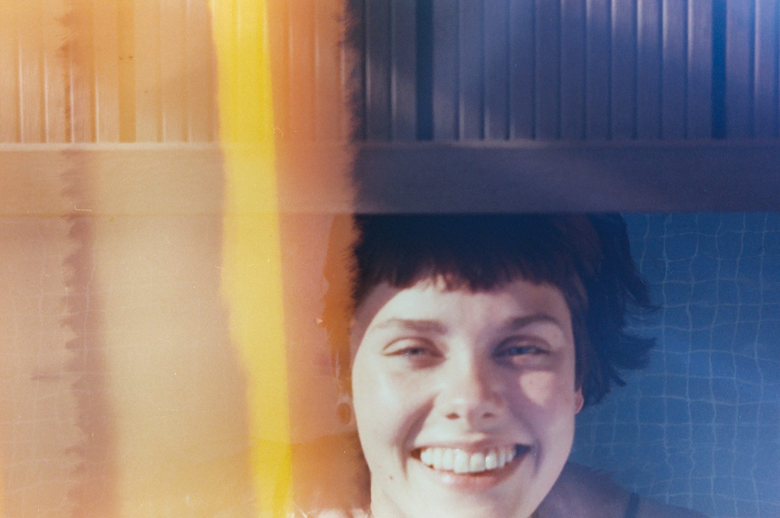 Analogue photograph of a young woman laughing by a swimming pool, one side flooded with orange light leaks from expired Fujicolor 200 film, the other showing turquoise pool water. A Hard Night Day by Eva Contreras, Woman Portrait Gallery.
