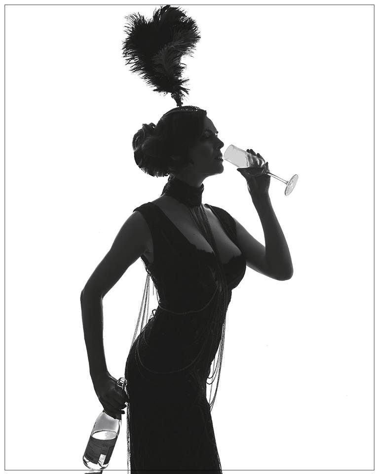High-contrast photograph of a woman in a feathered burlesque costume drinking champagne, rendered almost as a silhouette against a bright overexposed white background. La Femme Burlesque by Zora Jurenkova, Woman Portrait Gallery.