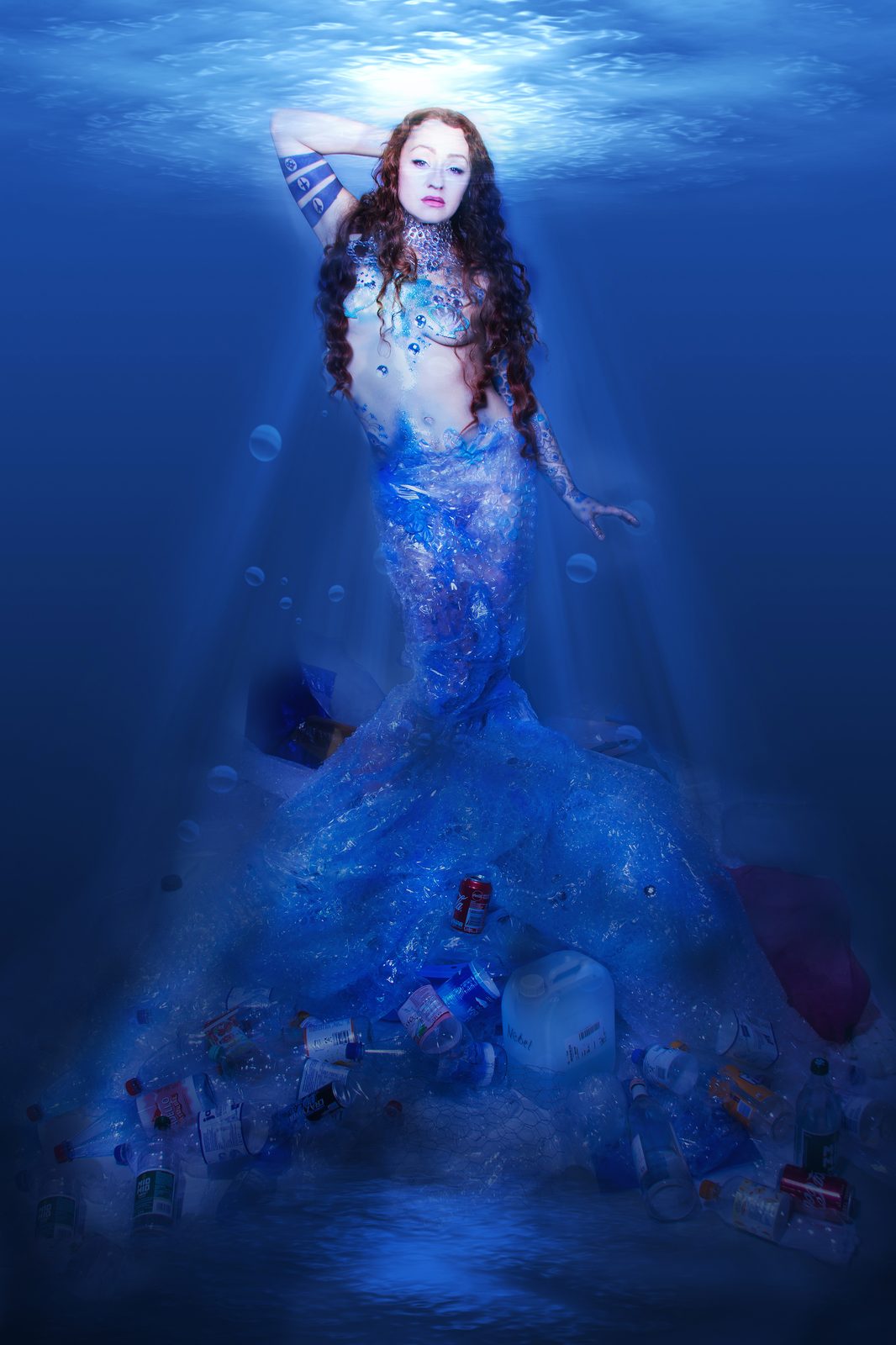 Digitally manipulated underwater photograph of a mermaid figure ascending, her flowing tulle-like fishtail trailing downwards revealing ocean floor debris. A commentary on ocean pollution. Venus the mermaids prayer by Zora Jurenkova, Woman Portrait Gallery.